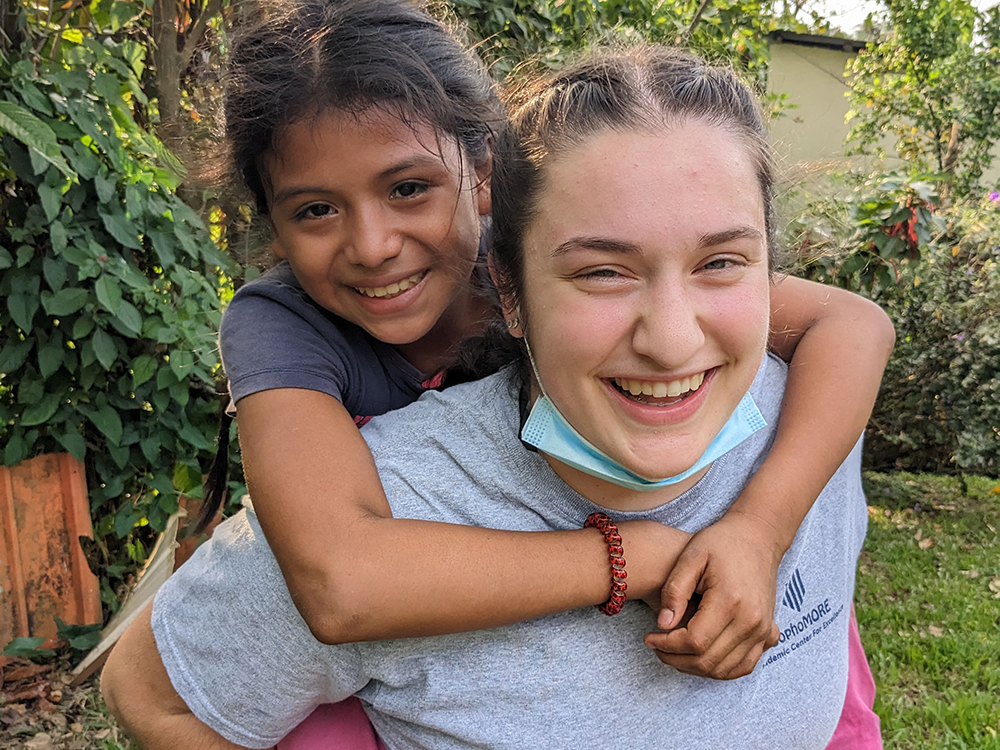 Two smiling girls outdoors, with the younger one piggybacking on the older one. The older girl has a face mask hanging below her chin. They are surrounded by greenery, capturing a joyful moment during their Guatemala service trip. Saint Joseph's College of Maine