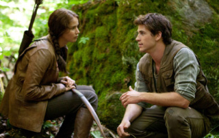 What We Talk About When We Talk About The Hunger Games 4 A woman holding a bow and arrows sits facing a man crouching in a forest, engaging in what appears to be an intense book discussion. The scene mirrors an encounter from The Hunger Games, blending reality with literature analysis. Saint Joseph's College of Maine