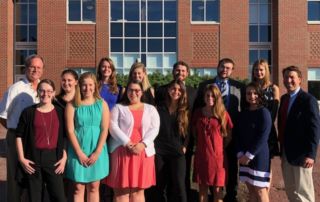 Delta Epsilon Sigma: Honoring Student Achievement 1 A group of 14 people, dressed in business and casual attire, stands outdoors in front of a brick building with large windows. They are posed in two rows, with some smiling at the camera, during a Delta Epsilon Sigma event honoring student achievement. Saint Joseph's College of Maine