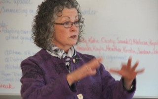 One Hundred Years Of Teaching Teachers 8 An older woman with curly gray hair wearing glasses and a purple blazer gestures with her hands while teaching in front of a whiteboard. Saint Joseph's College of Maine