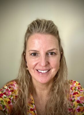 A woman with long, wavy blonde hair smiles at the camera in a colorful floral top, embodying the warmth and welcome of a counseling center against a plain light-colored background. Saint Joseph's College of Maine