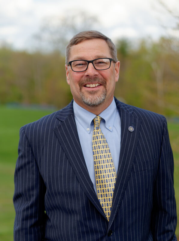 A man wearing glasses and a pinstriped suit with a yellow patterned tie stands outdoors on a grassy background. He has a goatee and is smiling at the camera, perhaps attending an inauguration ceremony. Saint Joseph's College of Maine