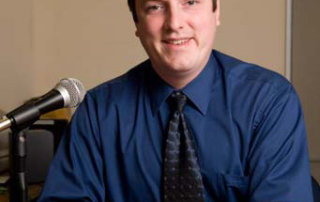 Justin Sparks '03 2 A man in a blue shirt and dark tie sits at a desk with a microphone in front of him, smiling at the camera in a neutral-toned office. This is Justin Sparks, class of 2003. Saint Joseph's College of Maine