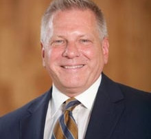 Meet Interim President Kenneth Lemanski 7 A man with short gray hair is smiling, wearing a dark suit, light shirt, and striped tie. The background is a blurred warm-toned setting. Kenneth Lemanski, Interim President, exudes confidence and professionalism. Saint Joseph's College of Maine