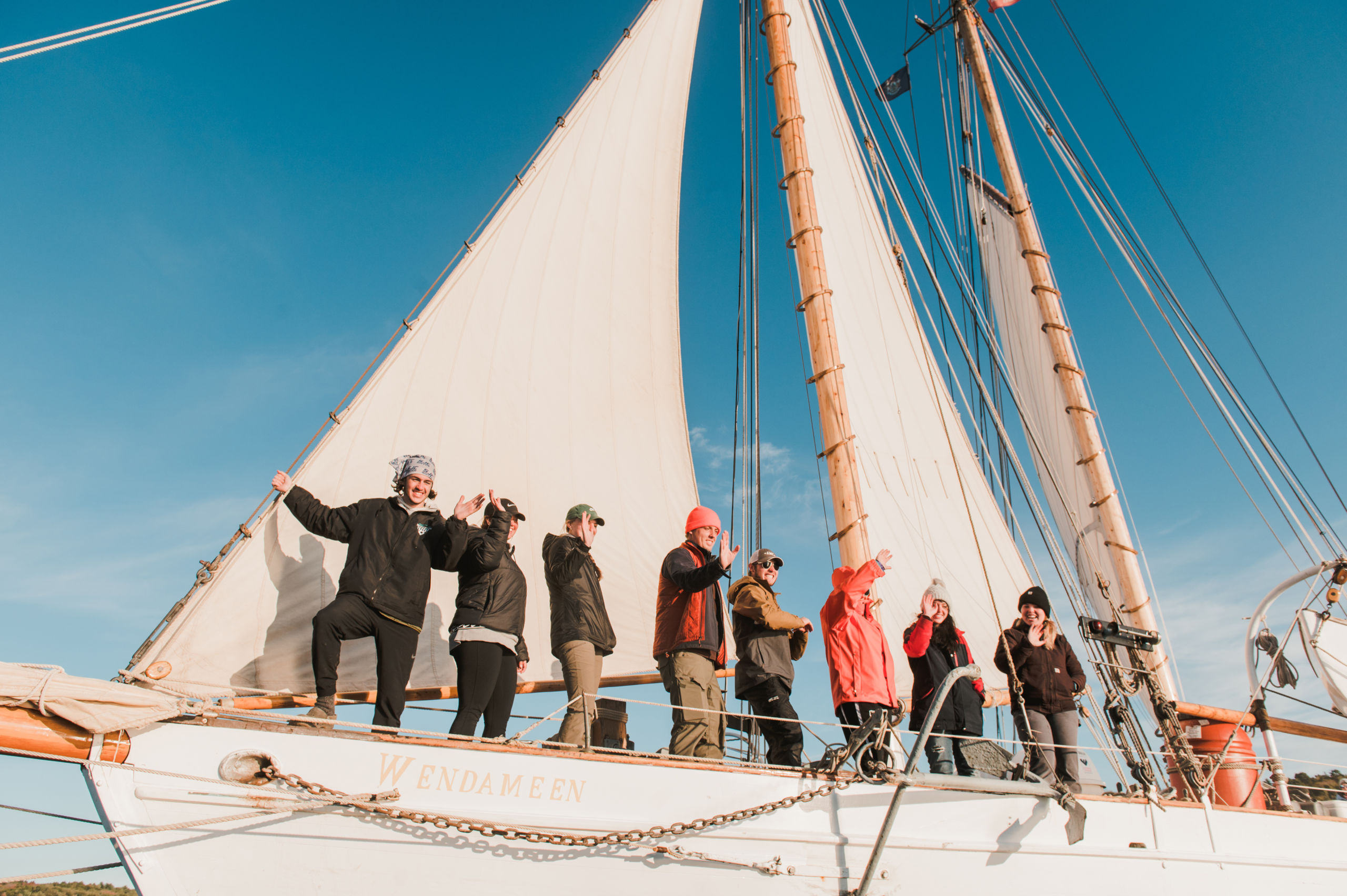 A group of people stand on the deck of a sailboat named WENDAMEEN, beneath the raised sails, on a sunny day. Some are waving and smiling while others look out to the horizon. Saint Joseph's College of Maine