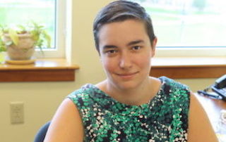 When Words Find Their Home 10 A person with short hair is sitting indoors by a window, wearing a sleeveless, patterned top. There is a plant on the windowsill and some office equipment on the desk, making it feel like home. Saint Joseph's College of Maine