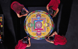 Tibetan Buddhist Monks Will Construct A Mandala Sand Painting And Perform Special Ceremonies At Saint Joseph’s College 9 Four Tibetan Buddhist monks in red robes meticulously craft an intricate sand mandala on a black surface at Saint Joseph’s College, viewed from above. Saint Joseph's College of Maine