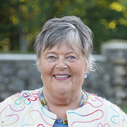 Center For Faith And Spirituality 9 A smiling older woman with short gray hair, wearing a light jacket with colorful swirls, stands outdoors with a blurred stone wall and greenery in the background. She exudes confidence as an esteemed member of the Board of Trustees. Saint Joseph's College of Maine