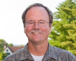 Professor Michael Connolly Receives Claddagh Award From Maine Irish Heritage Center 2 A man with glasses and a mustache, Professor Michael Connolly, smiles at the camera, standing outdoors with trees and houses in the background. Saint Joseph's College of Maine