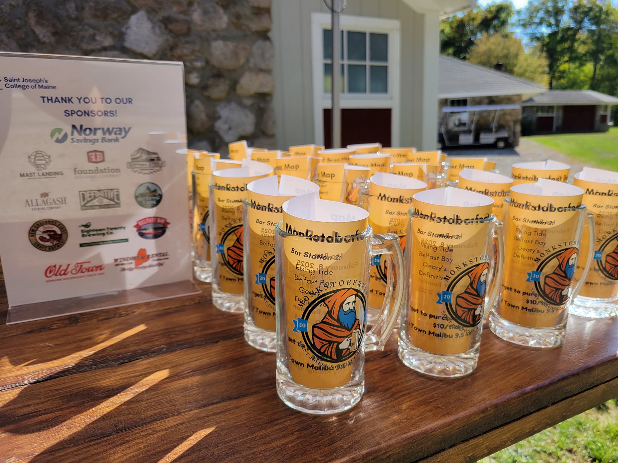 Alumni 3 A table displays multiple beer glasses with event logos. A sponsorship sign is visible on the left. The setting appears to be outdoors with buildings and greenery in the background. Saint Joseph's College of Maine