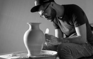 A man, wearing glasses and a cap, is focused on shaping a large pottery vase on a spinning wheel in an art workshop studio. Saint Joseph's College of Maine