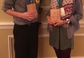 Emily Field And Nathan Morin Named Outstanding Future Professionals For Physical Education 8 Two people stand side by side, smiling and each holding award plaques for Outstanding Future Professionals. They wear business-casual attire with conference lanyards around their necks. The background features a beige wall and red carpeting, capturing the moment Emily Field is recognized for her achievements in Physical Education. Saint Joseph's College of Maine