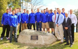 Plaque Dedication Ceremony Held For Clark’s Court 6 A group of people, most in matching blue jackets, are standing outdoors around a rock with a plaque on it. Some wear casual attire while others are dressed in business attire. The trees and clear sky provide a beautiful backdrop for the Plaque Dedication Ceremony at Clark’s Court. Saint Joseph's College of Maine