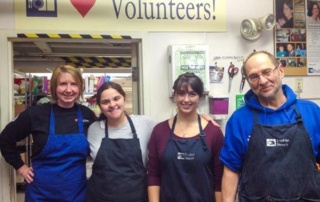 Students, Staff, And Faculty Volunteer At Preble Street Resource Center 5 Four people wearing aprons, including faculty volunteers and students, are standing in front of a "♥ Volunteers!" sign inside a room with various supplies and photographs on the walls. Saint Joseph's College of Maine