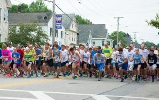 Westbrook Strong 5K Honors 2013 Alumnus, Endows Scholarship 1 A group of runners, wearing numbered bibs, starts the Westbrook Strong 5K race on a paved street in a residential area with houses and trees in the background. Saint Joseph's College of Maine