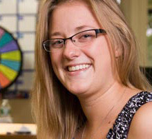 Faculty And Staff Mentors Help First-Year Students Ease Into Campus Life 10 A woman with long blonde hair, wearing glasses and a patterned sleeveless top, smiles at the camera. A colorful wheel and a calendar are visible in the blurred background, where she often offers guidance to first-year students. Saint Joseph's College of Maine