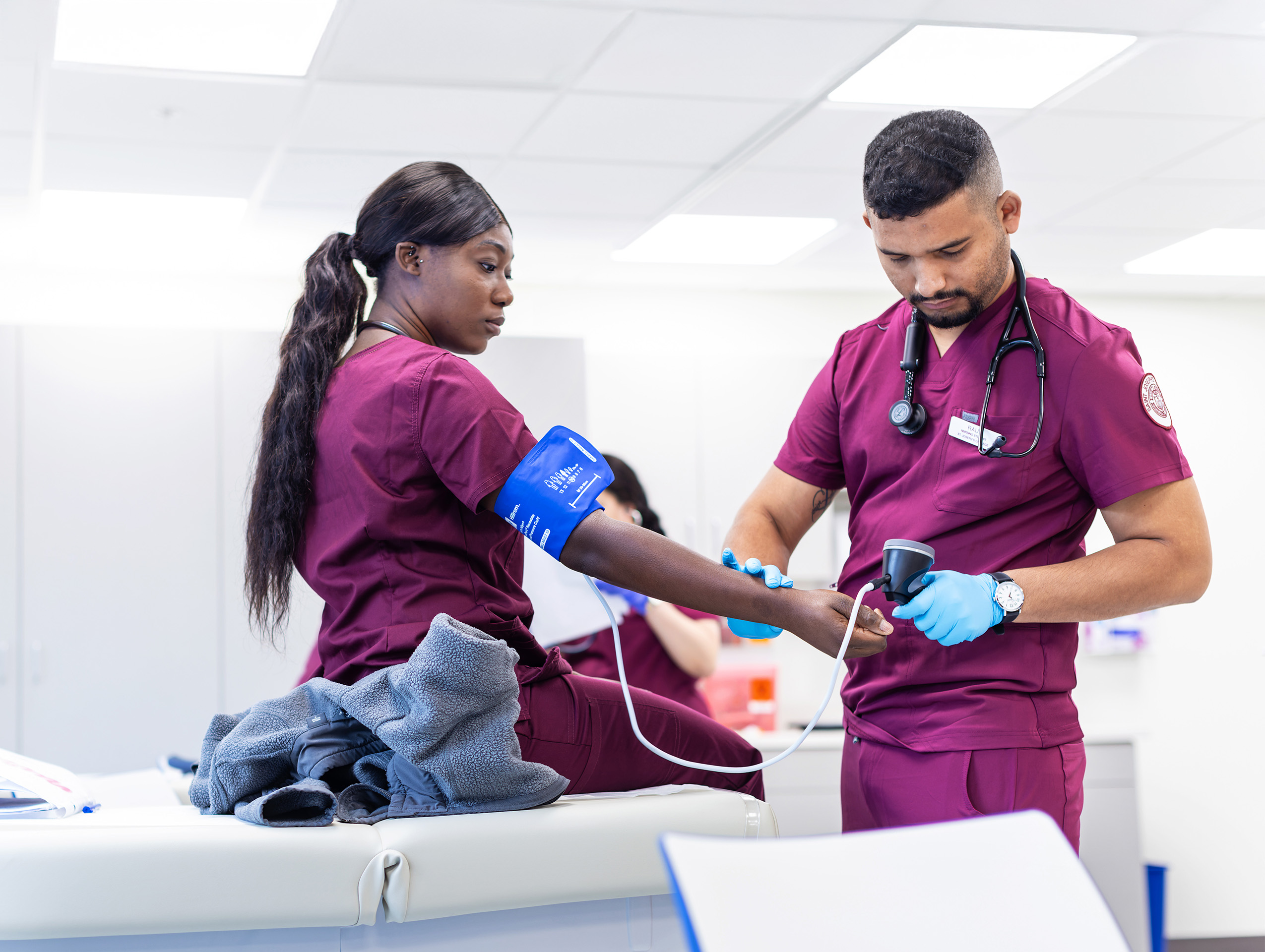 A healthcare worker in maroon scrubs checks a patients blood pressure in a clinical setting. Saint Joseph's College of Maine