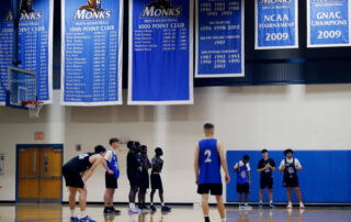 Basketball players practice on a court under blue banners displaying team achievements, including highlights from the NCAA tournament. Saint Joseph's College of Maine