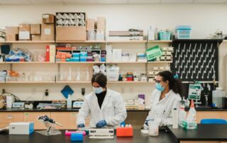 Two scientists wearing lab coats and protective masks work in a laboratory resembling those found at Saint Joseph's College of Maine. The lab, equipped with various scientific instruments and supplies on shelves and countertops, reflects the high standards of the Doctor of Chiropractic Program. Saint Joseph's College of Maine