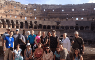 Study In Italy Or Spain This Summer! 2 A group of people posing for a photo inside the Colosseum in Rome, with the ancient stone structure visible in the background, during their summer abroad to study in Italy. Saint Joseph's College of Maine