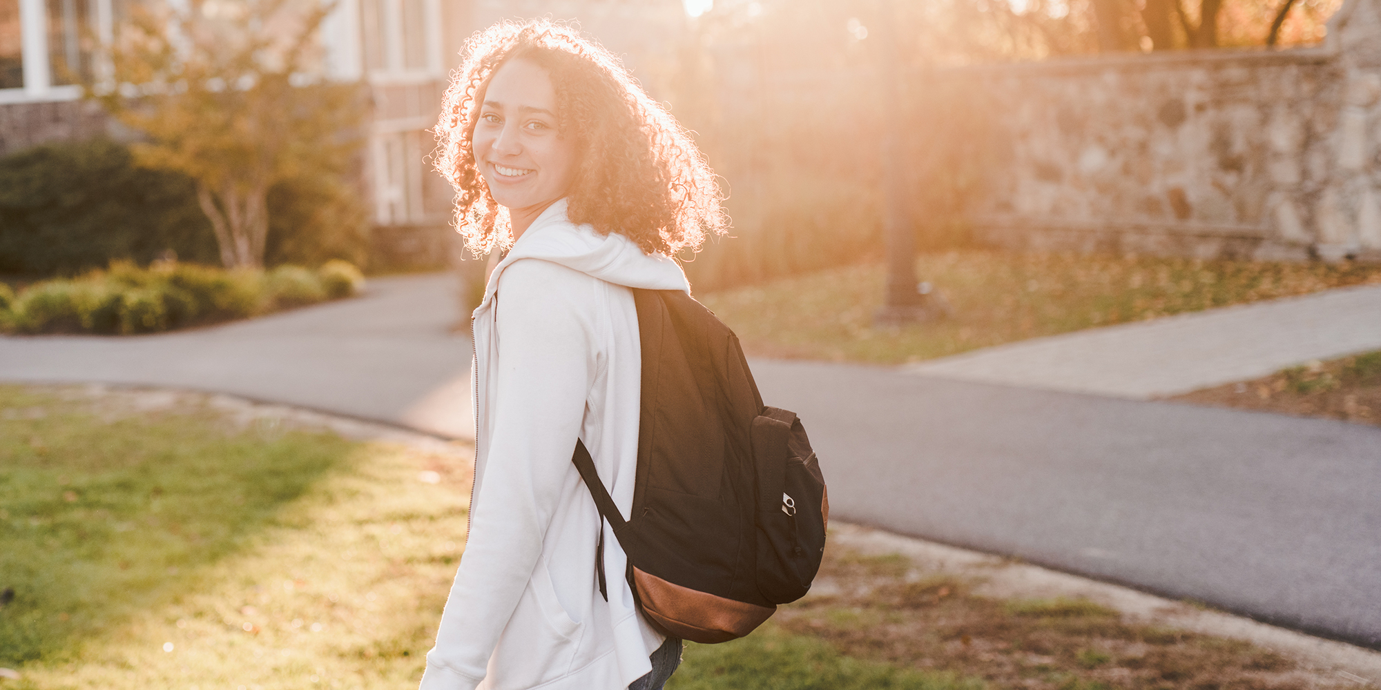 Person with curly hair and a backpack walking outdoors in a sunny park or campus setting, turning to look at the camera. Saint Joseph's College of Maine