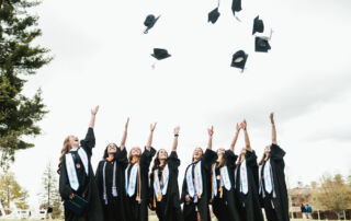 A group of SJC graduates in black gowns and caps toss their caps into the air outside, as the Class of 2024 joyously celebrates their graduation. Saint Joseph's College of Maine