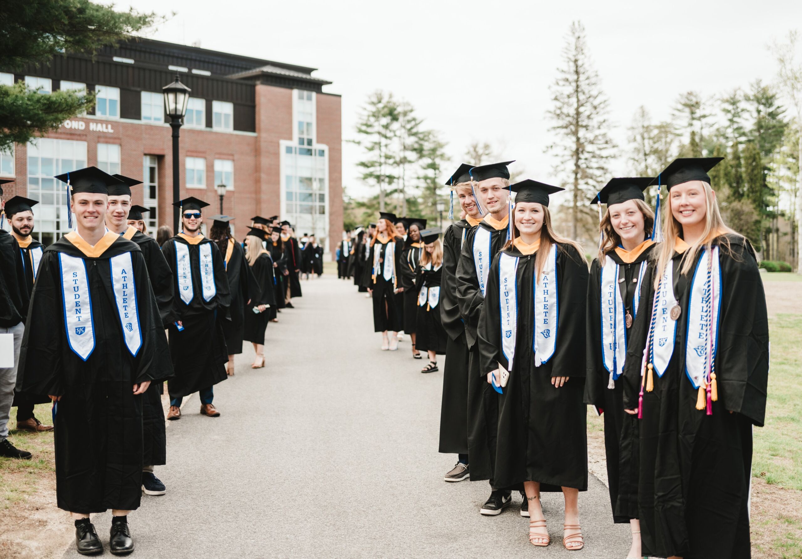 Commencement 2 A group of graduating students in caps and gowns stand in two lines on a campus path, celebrating commencement. Some are smiling and holding diplomas, with a building and trees in the background. Saint Joseph's College of Maine
