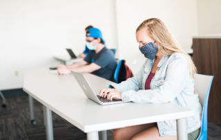 Three students wearing masks sit at desks, working on laptops in a bright classroom. The focus is on a student in a denim jacket in the foreground. Saint Joseph's College of Maine