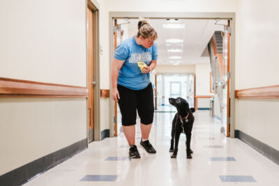 A woman wearing a blue shirt and black shorts walks down a hallway with a black dog, holding a yellow object in her hand. Saint Joseph's College of Maine