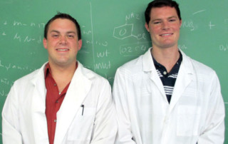 Students Conduct Research With Nasa In Mind 4 Two men in lab coats, engaged in student research, stand in front of a green chalkboard with scientific notations. Both are smiling and facing the camera. Saint Joseph's College of Maine