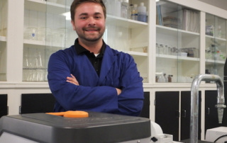 Erick Schadler ’18 Researches Infection Rates Of Deer Ticks In Raymond, Maine 2 A person wearing a blue lab coat, identified as Raymond Maine, stands with crossed arms in front of laboratory equipment and shelves containing glassware, likely studying infection rates caused by deer ticks. Saint Joseph's College of Maine