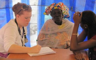 Students Afar: Senegal 9 A healthcare worker talks to two women under a blue tent. One woman, dressed in colorful traditional attire from Senegal, listens attentively while the other, possibly student Afar with long braided hair, rests her chin on her hand. Saint Joseph's College of Maine
