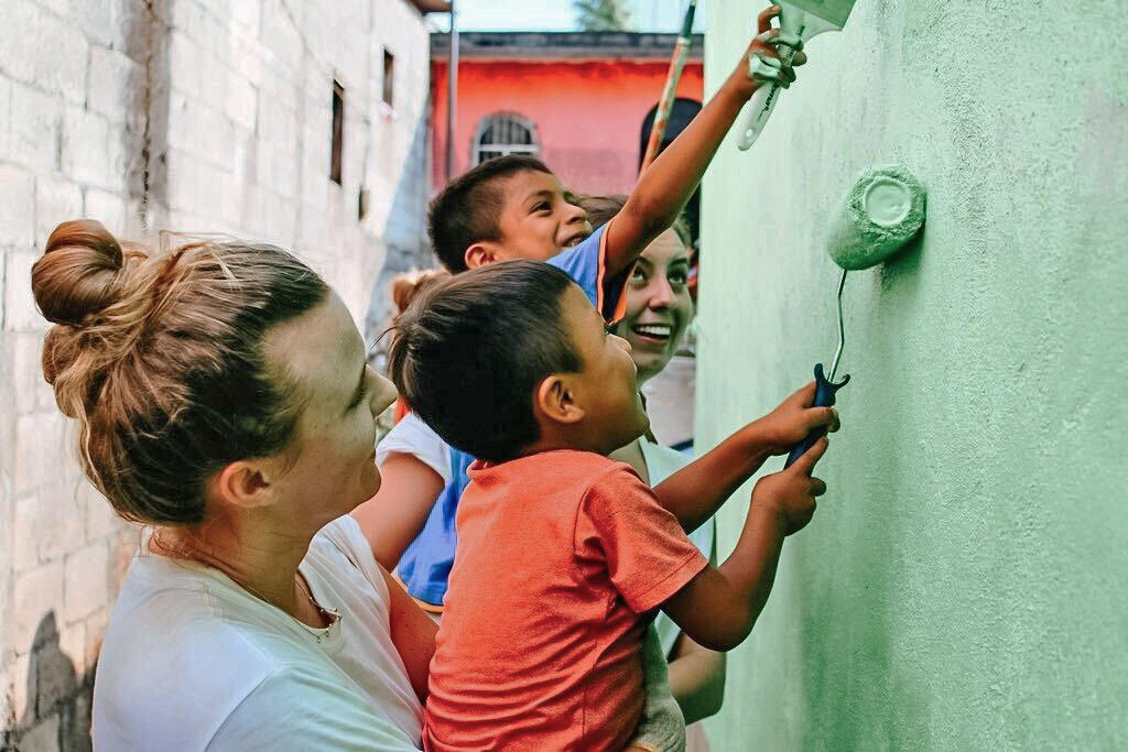 Servicetrips_Aprilquote Three adults and two children are painting a wall green together. One child uses a roller while another holds a paintbrush. They appear to be in an outdoor setting. Saint Joseph's College of Maine