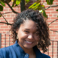 Experience Sjc 1 A woman with curly hair wearing a blue jacket stands outside in front of a brick wall and a tree, capturing the serene environment as she experiences Saint Joseph's College of Maine campus. Saint Joseph's College of Maine