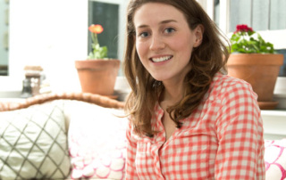 Healing At Camp Kita 1 A woman with long brown hair is sitting indoors on a patterned sofa, smiling. She is wearing a red and white checkered shirt. There are potted plants in the background, adding to the healing ambiance reminiscent of Camp Kita. Saint Joseph's College of Maine