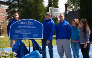 At Its Core, Clark’s Court Celebrates Students 5 A group of students, some in blue jackets, stand around a blue sign that reads "Clark's Court," as they celebrate on an outdoor basketball court. Saint Joseph's College of Maine