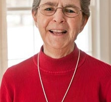 Spring Break Workfest Turns 20 2 Elderly woman with short gray hair and glasses, wearing a red sweater and a silver cross necklace, smiling in front of a window decorated for Spring Break. Saint Joseph's College of Maine