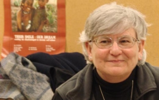 Alumna’s Work With Hispanic Community In Maine Is Her Calling 3 A person with gray hair and glasses sits in front of a poster and a brown backdrop, wearing a black top and a gray vest, reflecting the dedication to the Hispanic community that is evident in this distinguished alumna's work. Saint Joseph's College of Maine