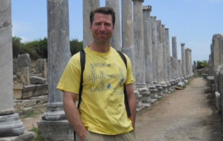 The Academy Of Travel: What You Learn Can Surprise 9 A man in a yellow T-shirt and khaki shorts stands between two rows of ancient stone columns on a dirt path, with a backpack strap visible over his shoulders. As part of The Academy of Travel, he's discovering history firsthand beneath a clear blue sky. Saint Joseph's College of Maine