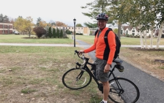 Saint Joseph’s College Earns Sustainable Commuting Award For Way 2 Go Maine Challenge 4 A person wearing an orange jacket and helmet stands next to a black bicycle on a path in a residential area, proudly displaying their Sustainable Commuting Award from Saint Joseph’s College as part of the Way 2 GO MAINE Challenge. Saint Joseph's College of Maine