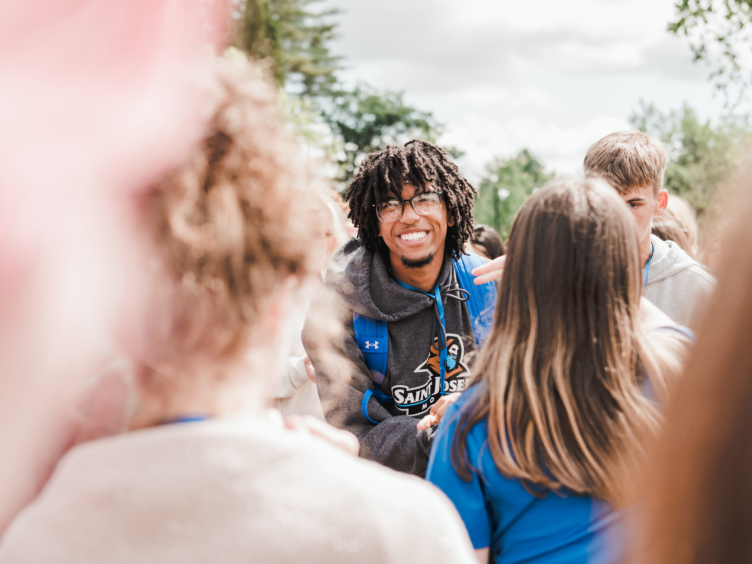 Student Life 1 A person with dreadlocks and glasses smiles while standing in the middle of a group of people outdoors on a sunny day, capturing the essence of student life. Saint Joseph's College of Maine