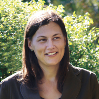Student_Angie Gleason A woman with shoulder-length brown hair stands in front of green bushes, smiling slightly. She is wearing a dark jacket and a black top, proudly holding her history-political science degree. Saint Joseph's College of Maine