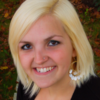 Student_Chelsea Candage A woman with a bachelor in history, short, straight blond hair, wearing large gold earrings and a black top, smiles at the camera. Saint Joseph's College of Maine