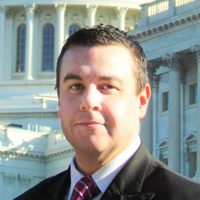 Student_Jeremiah Cottle A man with a history-political science degree stands in front of a large white building with columns and arched windows, wearing a suit and red tie. Saint Joseph's College of Maine