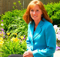 Tamera Edison ’10 4 A woman with red hair wearing a blue blazer, Tamera Edison, sits in front of a garden adorned with colorful flowers and lush green plants. An alumni of her university, she reflects on her graduation day amidst the vibrant backdrop. Saint Joseph's College of Maine