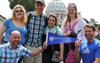 The Rome Experience 8 A group of six people are smiling and holding up a "Saint Joseph's College" pennant in an outdoor setting with a domed building in the background, celebrating their Rome Experience. Saint Joseph's College of Maine
