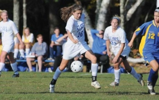 Getting Into Game Day 8 A soccer player in a white uniform prepares to kick a ball while other players in white and blue uniforms look on from the field. In the background, spectators are seated, observing the sports event on game day. Saint Joseph's College of Maine