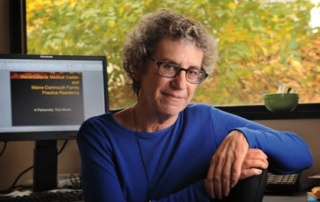 Nancy Weingarten '96, Msha, Keeps A Residency Running 2 Nancy Weingarten, a person with curly gray hair and glasses, sits at a desk in an office. Behind her is a computer displaying text and a large window with a view of trees, suggesting her residency at MSHA is both peaceful and productive. Saint Joseph's College of Maine