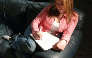 &Quot;Write Me A Letter.&Quot; 8 A person with long hair and a red shirt is sitting on a black leather couch, writing a letter in a notebook placed on their lap. Saint Joseph's College of Maine