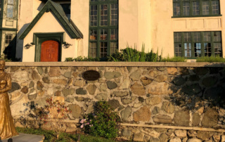 A bronze statue stands in front of a stone wall and a historic building with large windows and a wooden door, capturing the essence of a mission from days gone by. Saint Joseph's College of Maine